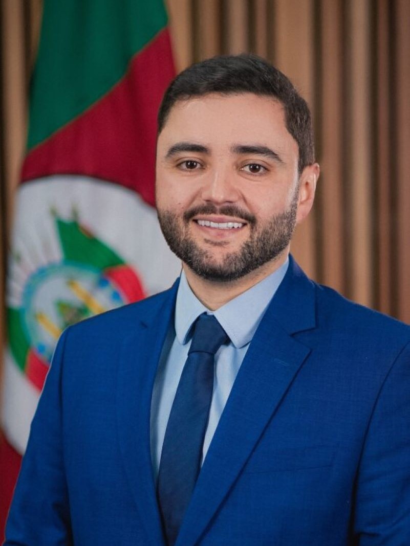 Retrato do Vice-governador Gabriel Souza, de terno azul com a bandeira do Rio Grande do Sul ao fundo esquerdo.