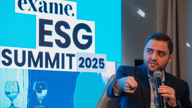 A imagem mostra o vice-governador do Estado, Gabriel Souza, em um painel de discussão. Ele é um homem branco de barba curta, vestindo um terno azul-escuro e camisa clara, sentado em uma poltrona cinza. Com uma mão, ele segura um microfone e com a outra, aponta para frente enquanto fala, com a boca ligeiramente aberta. Ao fundo, um telão azul exibe em branco o logotipo "exame." e as palavras "ESG SUMMIT 2025" em letras grandes. Dois copos de água vazios estão em uma mesa de vidro à frente do telão. Gabriel Souza foi um dos painelistas do evento ESG Summit 2025, que discute o futuro do clima e é promovido pela revista Exame, participando do painel "Eventos climáticos extremos e a necessidade de adaptação".
