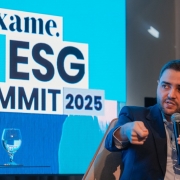 A imagem mostra o vice-governador do Estado, Gabriel Souza, em um painel de discussão. Ele é um homem branco de barba curta, vestindo um terno azul-escuro e camisa clara, sentado em uma poltrona cinza. Com uma mão, ele segura um microfone e com a outra, aponta para frente enquanto fala, com a boca ligeiramente aberta. Ao fundo, um telão azul exibe em branco o logotipo "exame." e as palavras "ESG SUMMIT 2025" em letras grandes. Dois copos de água vazios estão em uma mesa de vidro à frente do telão. Gabriel Souza foi um dos painelistas do evento ESG Summit 2025, que discute o futuro do clima e é promovido pela revista Exame, participando do painel "Eventos climáticos extremos e a necessidade de adaptação".