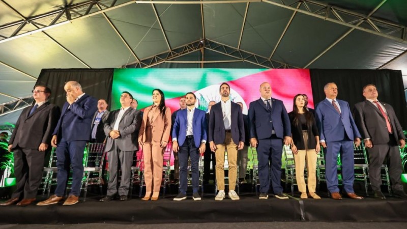 Imagem de uma cerim&ocirc;nia oficial em um palco coberto por tenda. Cerca de doze pessoas adultas est&atilde;o em p&eacute;, lado a lado, usando trajes formais. Ao fundo, h&aacute; cadeiras transparentes organizadas em fileiras e um grande painel de LED com faixas nas cores verde, branco e vermelho. A ilumina&ccedil;&atilde;o &eacute; forte e o ambiente transmite formalidade institucional.