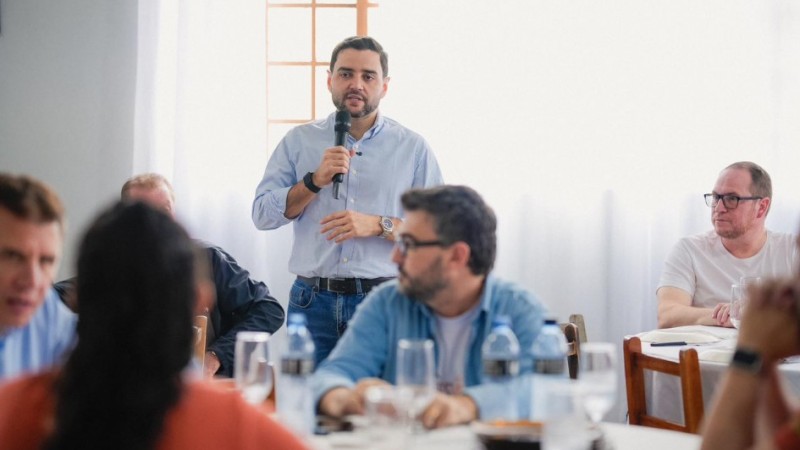 Imagem de um encontro em ambiente interno e bem iluminado. Ao centro, o vice-governador Gabriel Souza est&aacute; em p&eacute; segurando um microfone e falando. Ao redor, outras pessoas est&atilde;o sentadas em mesas com copos e garrafas de &aacute;gua, aparentando ouvir. O espa&ccedil;o lembra uma reuni&atilde;o ou conversa coletiva.