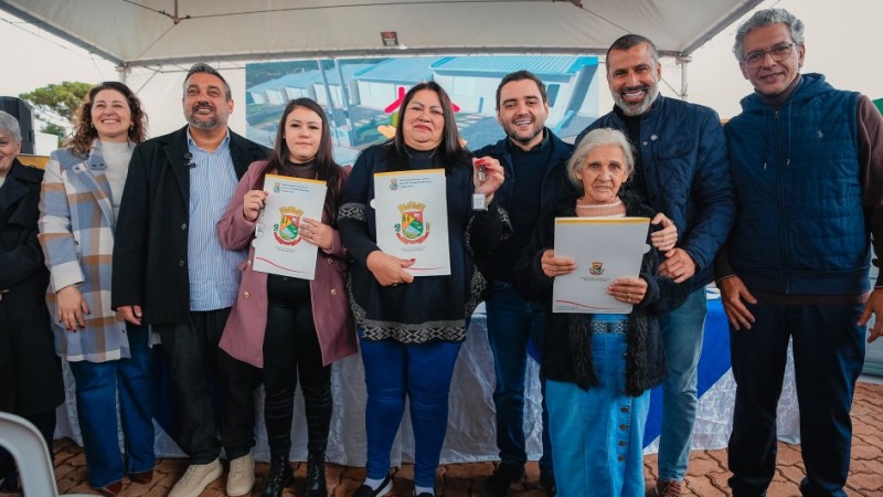 Grupo de oito pessoas posando sob uma tenda em &aacute;rea externa. Algumas seguram documentos com bras&atilde;o e uma pessoa exibe uma chave, sugerindo uma entrega oficial. Ao fundo, h&aacute; uma mesa com toalha azul e branca e um painel com imagem de empreendimento.