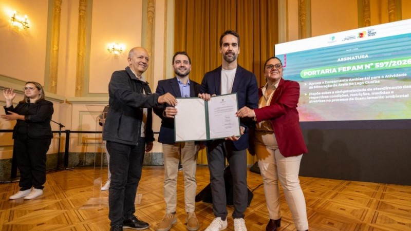 Quatro pessoas, entre elas o governador Eduardo Leite e o vice-governador Gabriel Souza, posam no palco de um sal&atilde;o institucional, segurando um documento aberto durante um ato de assinatura oficial; ao fundo, h&aacute; um tel&atilde;o com texto sobre uma portaria ambiental e, &agrave; esquerda, uma int&eacute;rprete de Libras.