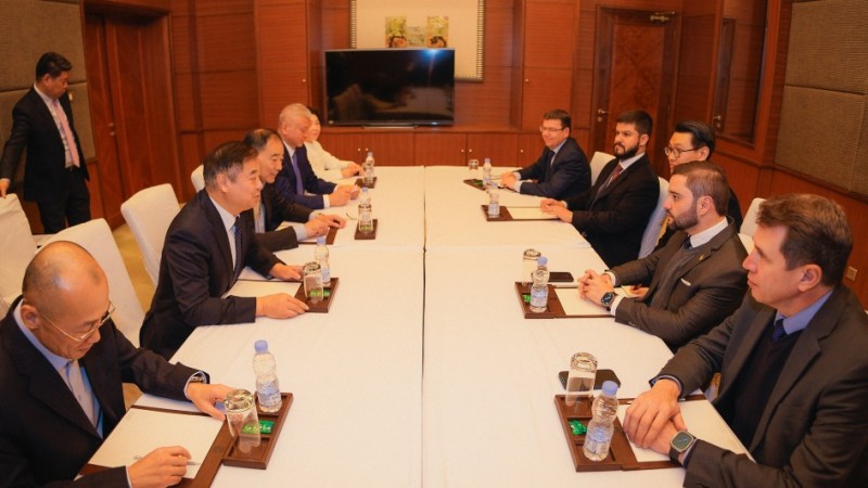 Reuni&atilde;o formal em sala corporativa com mesa comprida ao centro, onde duas fileiras de pessoas de terno est&atilde;o sentadas frente a frente. Sobre a mesa h&aacute; garrafas de &aacute;gua, copos e celulares. Ao fundo, uma TV desligada e portas fechadas nas laterais. Uma pessoa permanece em p&eacute; no canto esquerdo, observando.